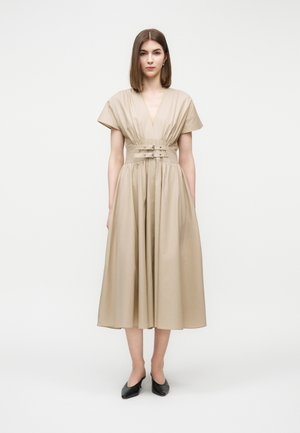 Žena oblečená v béžových midi šatách s krátkymi rukávmi, hlbokým výstrihom do V, opaskom v páse a čiernymi nízkymi opätkami, stojaca pred jednoduchým pozadím.
