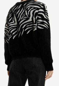 Schwarzer Pullover mit flauschiger Textur, der auf der Rückseite ein weißes Zebramuster aufweist. Kombiniert mit hochgeschnittenen schwarzen Jeans.