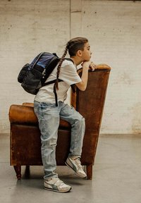 Een kind in een wit T-shirt, versleten blauwe jeans en grijze schoenen ligt op een bruine leren fauteuil, met een zwart en blauwe rugzak.