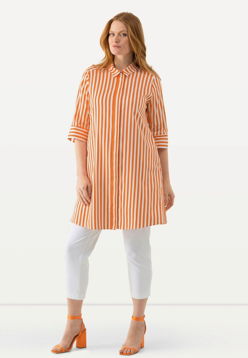 Camicia tunica a righe arancioni e bianchi con maniche 3/4, abbinata a pantaloni bianchi cropped e sandali con tacco a blocco arancioni.