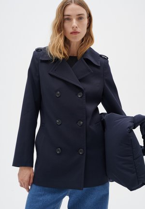 Navy double-breasted coat with large buttons and shoulder details, worn over a black ribbed sweater, with a puffy navy bag in hand.
