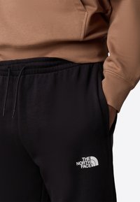 Zwarte joggers met elastische tailleband en trekkoord, met een wit logo op de linker bovenbeen. Gepaard met een bruine hoodie.