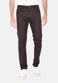 Pantaloni slim-fit marroni realizzati in misto cotone, con chiusura a bottone, tasche frontali e posteriori, e una texture liscia. Abbinati a sneaker bianche.