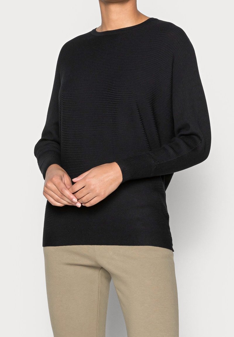 Personne portant un pull côtelé à manches longues noir avec un pantalon beige, debout devant un fond clair uni.