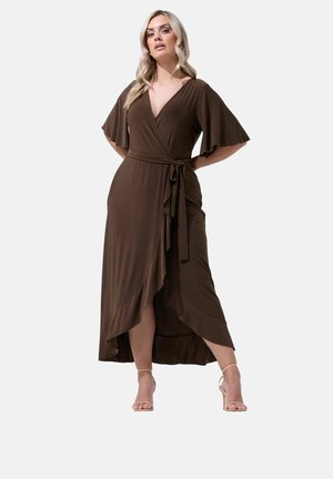 Vrouw in een bruine wikkeljurk met wapperende mouwen en een voorstrik, gecombineerd met naakte hoge hakken, staand tegen een effen achtergrond.