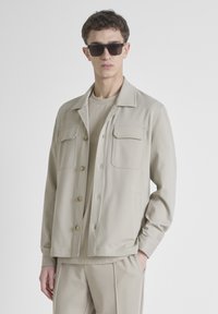 Beige jas met twee borstzakken en knoopsluiting, gecombineerd met een bijpassend lichtkleurig topje, tentoongesteld met een zonnebril.