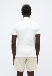 Λευκό πόλο πουκάμισο με κλασικό γιακά, κοντά μανίκια και διακριτική αντίθετη λεπτομέρεια. Συνδυασμένο με ανοιχτού μπεζ χρώματος σορτς. Απαλή υφή από βαμβάκι.