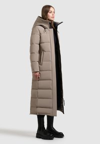 Abrigo acolchado beige de diseño largo, con un gran cuello, forma cuadrada, patrón de canalé vertical y bolsillos laterales. Se lleva con botas negras gruesas.