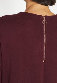 Dettaglio della parte superiore della schiena di una persona che indossa un maglione bordeaux con una zip verticale dorata e un tab circolare al collo.