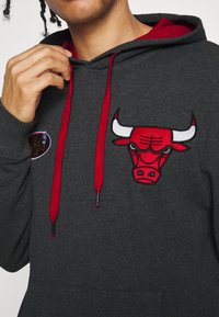 Sudadera gris con cordones y forro rojos, que presenta un destacado gráfico bordado de un toro en rojo y blanco y un parche de logo en el pecho.