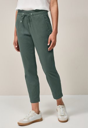 Pantaloni tapered in misto cotone verde con vita elasticizzata e cordoncino. Presentano tasche laterali e una texture liscia. Abbinati a sneakers bianche.
