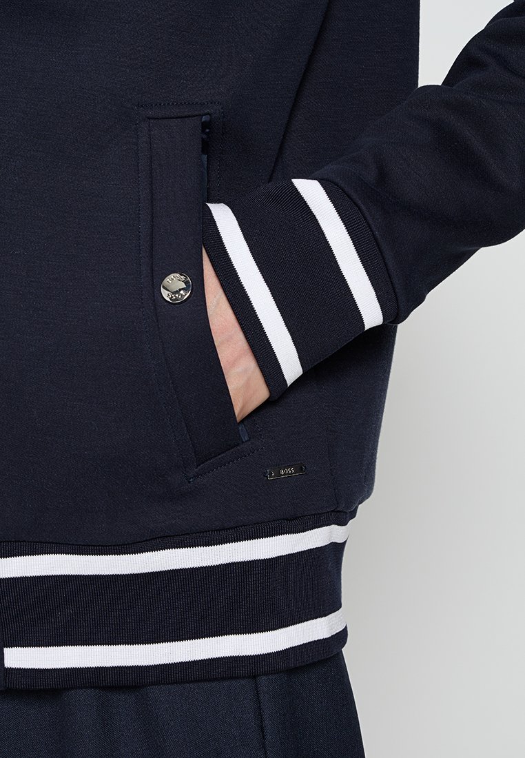 Granatowy sweter z białymi prążkowanymi mankietami, z boczną kieszenią i małą metką z logo przy dolnym brzegu. Miękka struktura materiału.