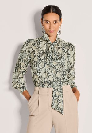 Green snake-print blouse with a tie collar, long sleeves, gathered shoulders, and a semi-sheer texture, paired with high-waisted beige trousers.
