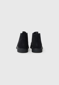 Botas de tobillo de ante negro con una suela de goma texturizada, que cuentan con lengüetas en la parte superior y costuras traseras lisas para un uso fácil. Diseño minimalista, sin herrajes visibles.
