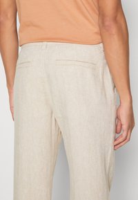 Ljust beige linbyxor med fiskbensmönster, som har två bakfickor och en avslappnad passform, visas från baksidan.