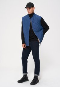 Nieprzemakalna, niebieska kamizelka pikowana z wysokim ko�łnierzem, zamkiem błyskawicznym z przodu i bez rękawów, zestawiona z czarnym dopasowanym golfem, ciemnymi dżinsami i sznurowanymi bootsami.