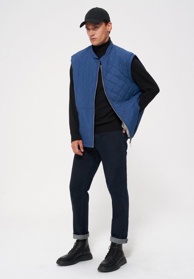 Nieprzemakalna, niebieska kamizelka pikowana z wysokim kołnierzem, zamkiem błyskawicznym z przodu i bez rękawów, zestawiona z czarnym dopasowanym golfem, ciemnymi dżinsami i sznurowanymi bootsami.