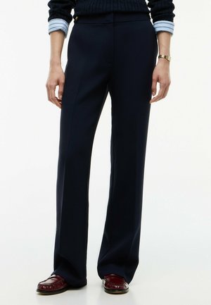 Person iført mørkeblå straight-leg bukser, brune loafers, en navy sweater med lyseblå skjorteflip og et guld armbåndsur.