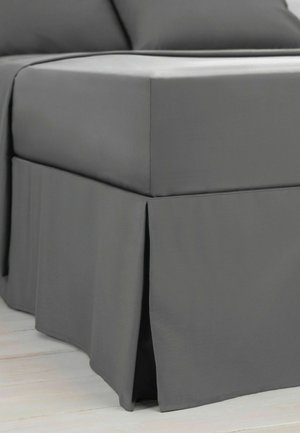 Nahaufnahme eines grauen Stoff-Bettrocks, der die Basis eines Bettes mit einem plissierten Eckdetail auf einem hellen Holzboden bedeckt.