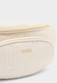 Creme læder crossbody taske med præget logo, guldramme detaljer og en rund form. Tekstureret overflade tilføjer visuelt interesse.