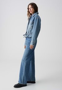 Spijkerjack met een koord in de taille, lichtblauwe kleur, en twee voorzakken, gecombineerd met wijde lichtblauwe jeans en zwarte laarzen.