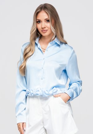 Femme aux longs cheveux blonds portant une chemise en satin bleu clair boutonnée et un pantalon blanc, debout avec une main dans la poche.