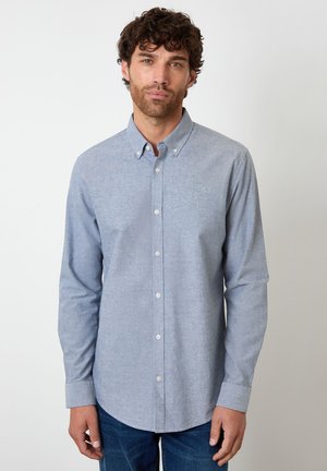 Uomo con capelli ricci che indossa una camicia azzurro chiaro con bottoni e jeans blu scuro, in piedi davanti a uno sfondo bianco semplice.