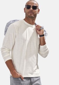 Helle beige Langarmbluse mit lockerer Passform, strukturiertem Stoff und Rundhalsausschnitt, kombiniert mit grauen Hosen und einer Sonnenbrille, die über der Schulter gehalten wird.