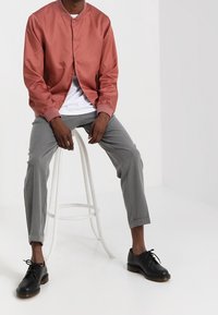 Man seated on white stool wearing a coral bomber jacket, white T-shirt, gray ankle-length pants, and black lace-up shoes.