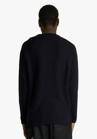 Persona con capelli corti e ricci che indossa un maglione a maglia semplice di colore blu scuro, vista di spalle su uno sfondo neutro.