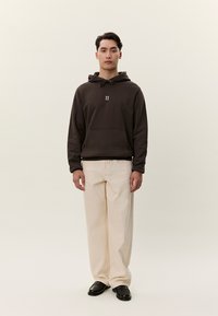 Giovane uomo con felpa con cappuccio marrone scuro con logo "II", pantaloni larghi color crema e scarpe nere, in piedi davanti a uno sfondo beige semplice.