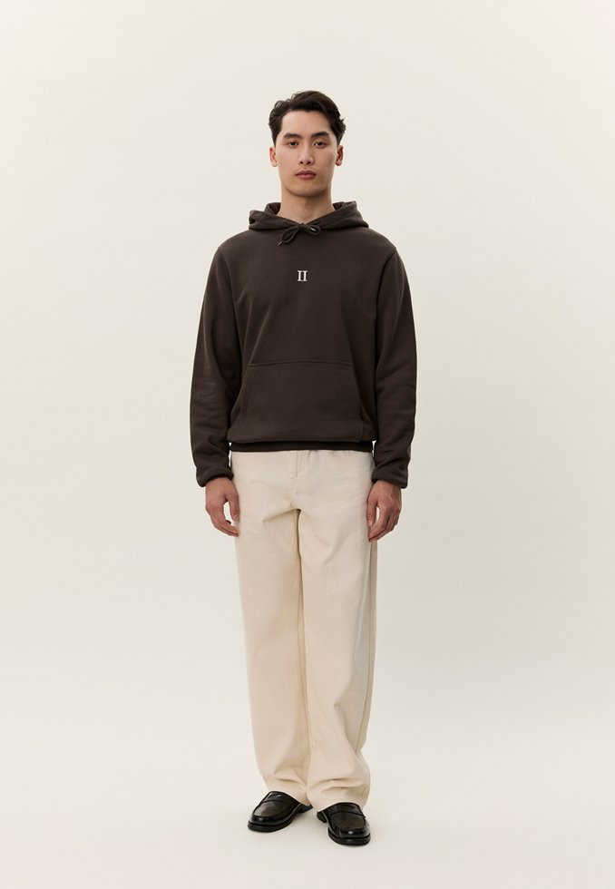 Giovane uomo con felpa con cappuccio marrone scuro con logo "II", pantaloni larghi color crema e scarpe nere, in piedi davanti a uno sfondo beige semplice.