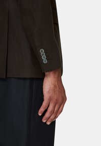 Blazer in lino marrone con vestibilità su misura, chiusura a singolo bottone e bottoni grigi sui polsini. Pantaloni neri visibili sotto.