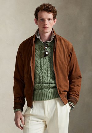 Mladý muž s kučeravými vlasmi oblečený v hnedej semišovej bunde, zelenom svetríku s vrkočovým vzorom, károvaných košeli, bielych nohaviciach a slnečných okuliaroch zavesených na golieri svetra.