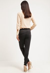 Beige blouse met lange mouwen gecombineerd met donkergrijze strakke broek en huidskleurige hoge hakken. De blouse heeft knoopdetails en de broek heeft achterzakken.