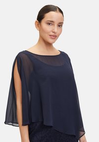 Navy modrý průhledný top s lodním výstřihem a volnými rukávy, vrstvený přes texturovaný krajkový spodní díl. Lehká tkanina, hladký povrch.