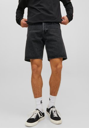Mand iført sorte denimshorts, hvide crew-sokker, sort-hvide sneakers og en sort langærmet skjorte, stående mod en ensfarvet grå baggrund.