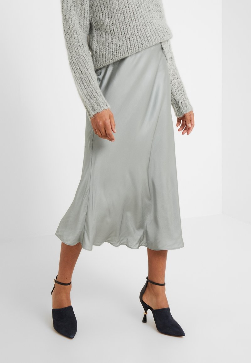 Femme portant une jupe midi en satin gris clair, un pull en maille gris et des escarpins noirs pointus à talons avec brides à la cheville, debout sur un sol blanc.