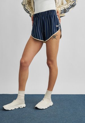 Person iført marineblå shorts med nålestriber, hvide sneakers og en stribet jakke med hænderne på hofterne, stående på en blå tæppe.