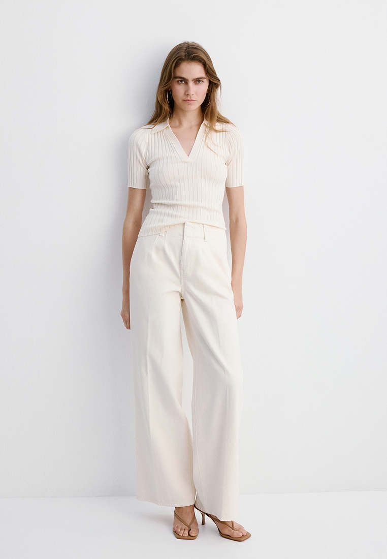 Femme debout contre un mur blanc portant un haut court côtelé crème à manches courtes, un pantalon large crème et des sandales à talons marron.