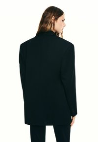 Hosszú barna hajú nő fekete, túlságosan nagy blézerben és hozzá illő fekete nadrágban, oldalt állva, fehér háttér előtt.