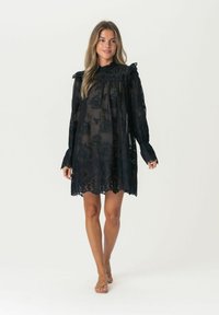 Schwarzes Spitzenkleid mit langen Ärmeln und einem gerüschten Kragen, das blumige Muster und eine leicht durchscheinende Textur aufweist, in knieumspielender Länge.