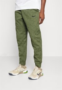 Calças jogger verdes de malha com punhos canelados, apresentando um logótipo Nike preto na coxa esquerda, combinadas com sapatilhas de desporto bege.