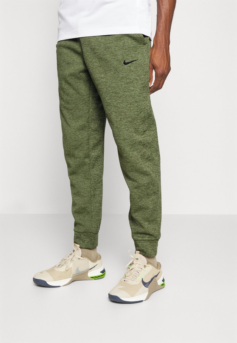 Calças jogger verdes de malha com punhos canelados, apresentando um logótipo Nike preto na coxa esquerda, combinadas com sapatilhas de desporto bege.