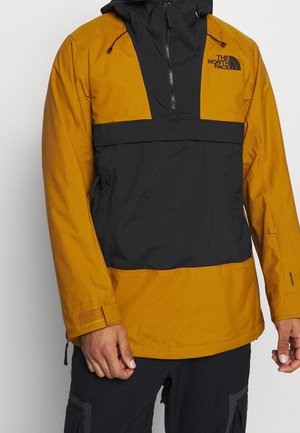 Homme portant une veste The North Face moutarde et noire avec une fermeture éclair partielle, une capuche et des sangles velcro aux poignets, debout devant un fond uni.