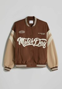 Brun varsityjacka med beiga ärmar, randiga manschetter och en vit grafisk text "Match Day" som tydligt syns. Har tryckknappar.