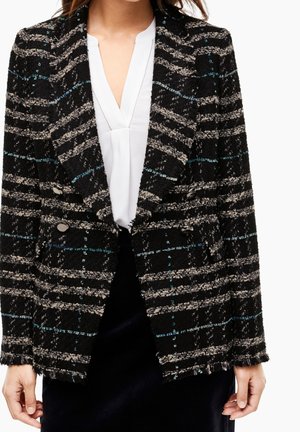 Schwarz-grau kariertes, strukturiertes, zweireihiges Sakko über einer weißen V-Ausschnitt-Bluse, getragen von einer Person mit locker an den Seiten hängenden Händen.