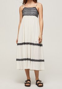Weißes Maxikleid mit schwarzem gesticktem Band an der Brust und dem Saum. Verfügt über verstellbare Träger und einen leichten, strukturierten Stoff. Schwarze Sandalen.