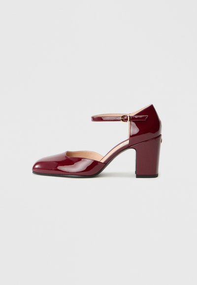 Chaussure en cuir verni bordeaux avec un bout pointu, une bride à la cheville et un talon bloc. Présente un design découpé sur les côtés.