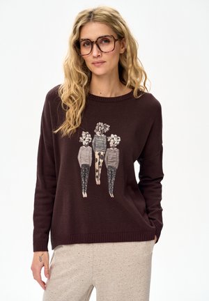 Blonde Frau mit großen Schildpattbrillen, einem braunen Pullover mit drei stilisierten Figuren mit Blumenköpfen und hellbeigen Hosen vor weißem Hintergrund.